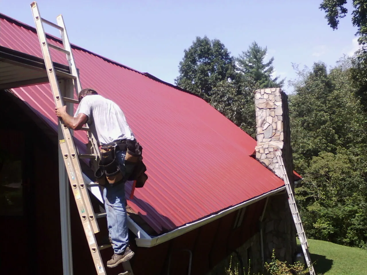 Roofer on a ladder beside a red metal roof and stone chimney — Soffit & Fascia Repair by Roofing Brothers in Columbus City